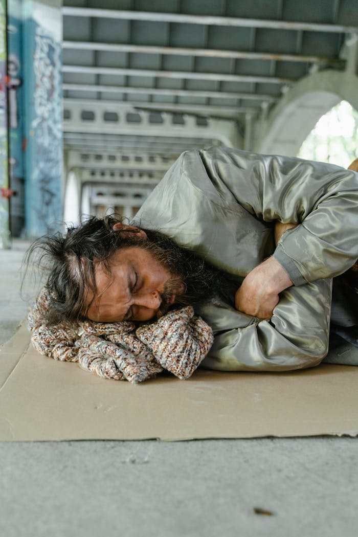 A homeless man with a beard is sleeping on a cardboard under a bridge, highlighting urban poverty.