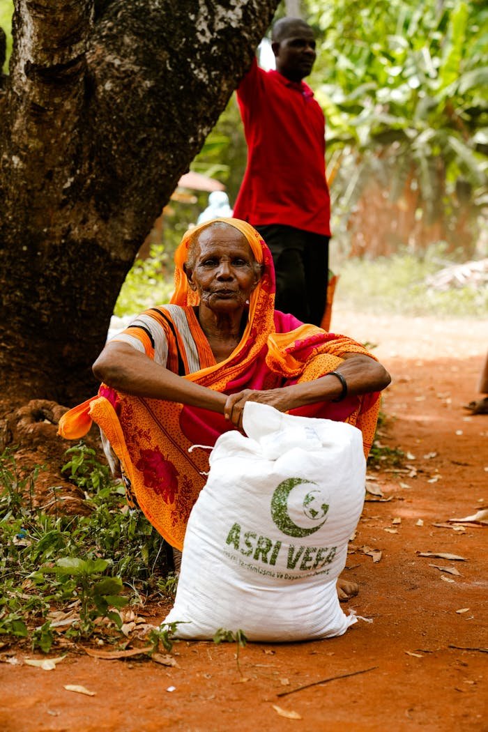 Elderly woman sits by tree with a relief bag labeled ASRI VEER. Rural outdoor setting.