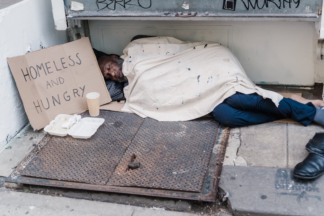 Street scene depicting a homeless man sleeping with a placard reading Homeless and Hungry.